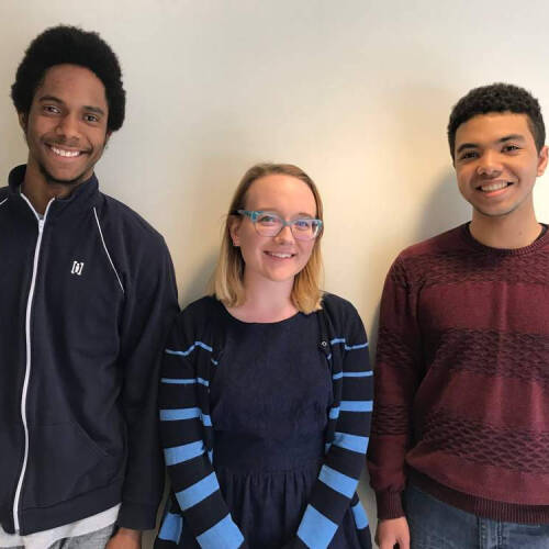 Three people stand against a gray wall. They are two boys and a girl in the middle.